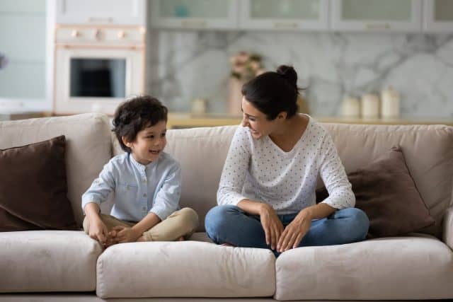 Happy Indian mother and adorable 5s son sitting on cozy couch together, smiling young mom with little son having fun, talking, spending enjoying leisure time weekend at home, laughing at joke