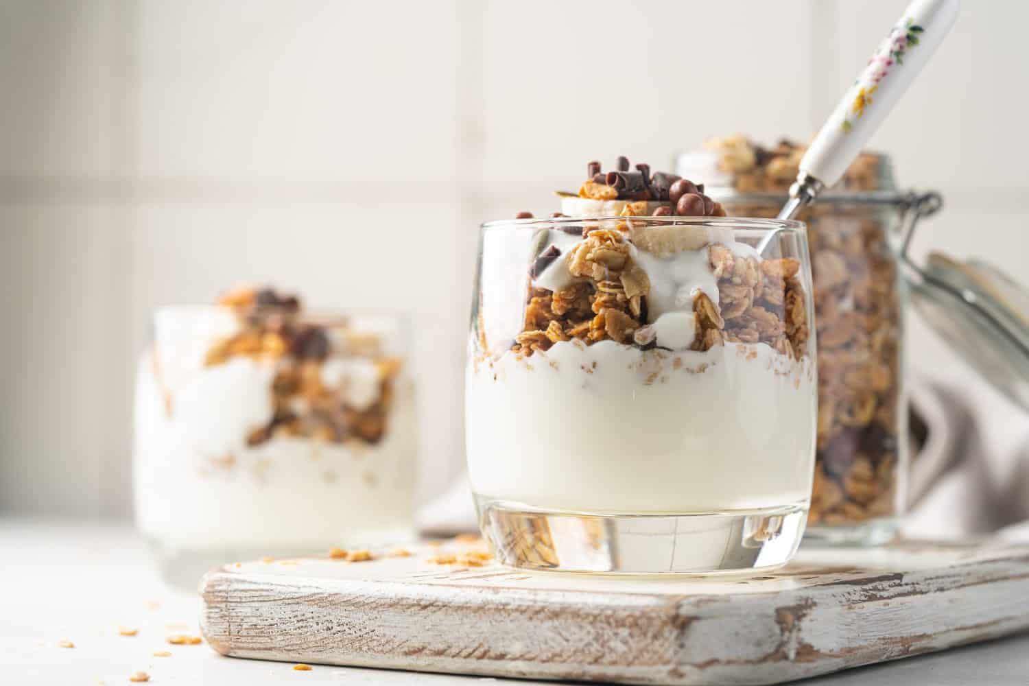 Crunchy granola with yogurt, banana, nuts, chocolate and honey in a glass on white background. Healthy breakfast concept.