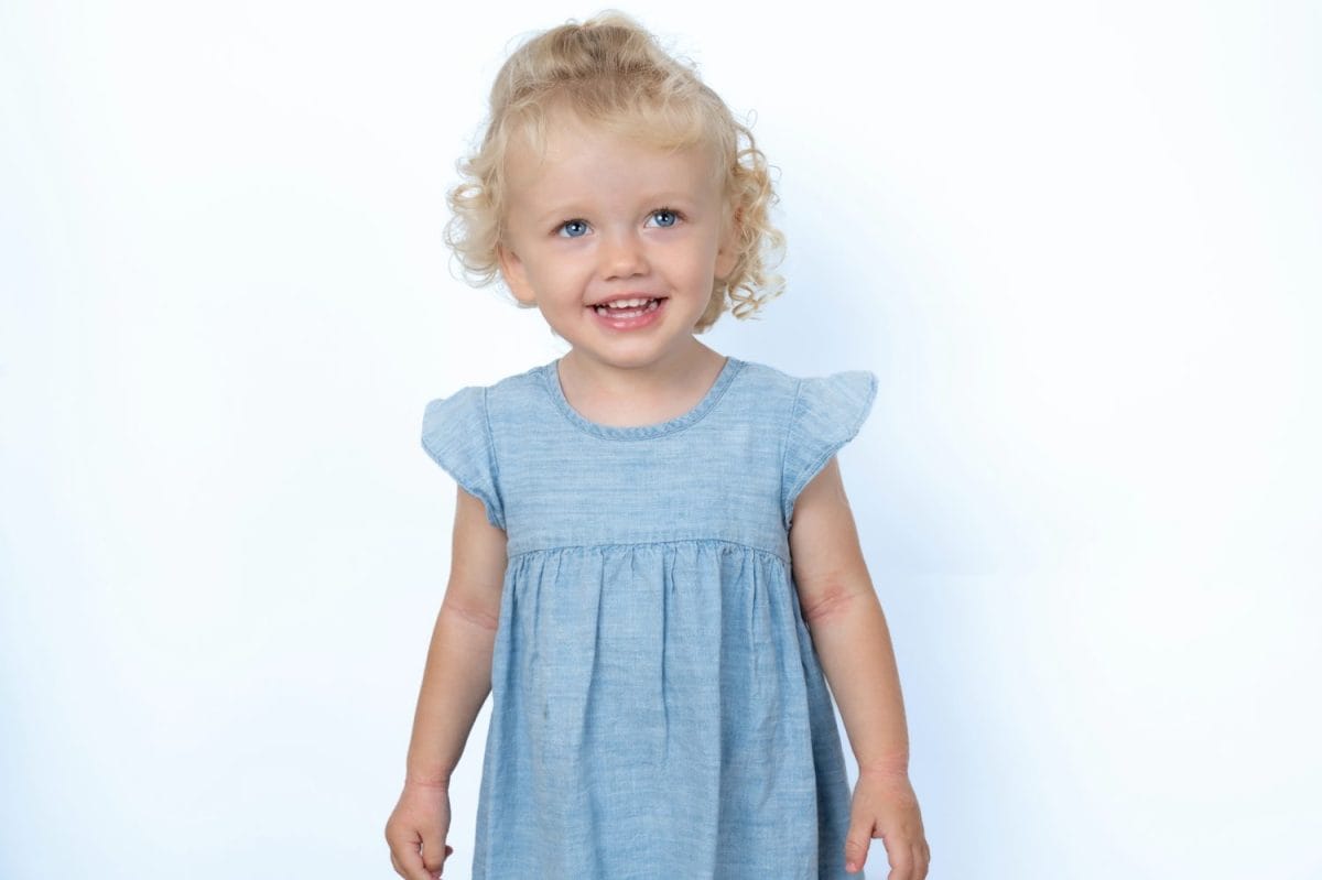 Beautiful baby girl wearing blue dress standing over white studio background smiling and looking aside. Childhood concept.