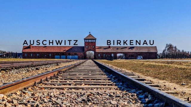 A train track is visible next to an industrial building with the entrance to the Auschwitz Birkenau concentration camp in the foreground