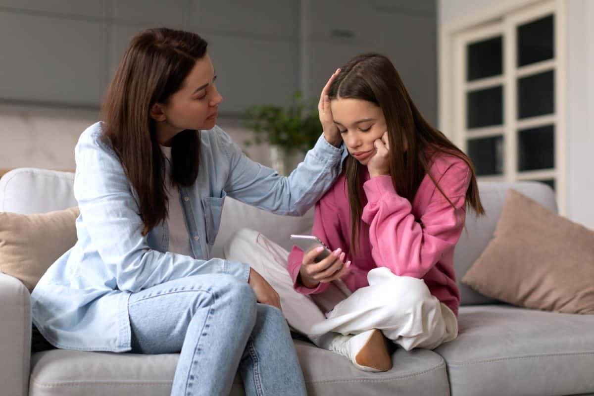 Supportive mother comforting sad teen girl rejected by friends, helping depressed daughter to deal with breakup, child looking at phone