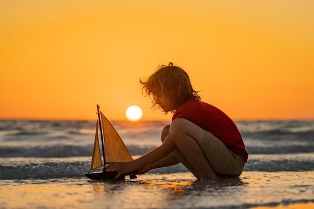 Little boy playing with toy sailing boat, toy ship. Travel and adventure concept. Child feeling adventurous while cruising. Kid on summer sunset beach. Kid playing on the beach at the sunset time.