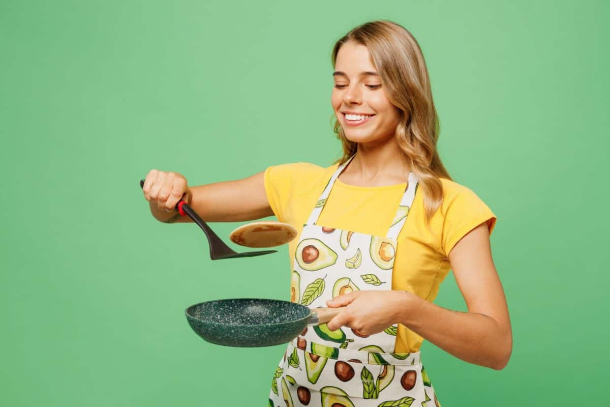 Young cheerful fun housewife housekeeper chef cook baker woman wear apron yellow t-shirt hold in hand pan spatula frying pancakes isolated on plain pastel green background studio. Cooking food concept