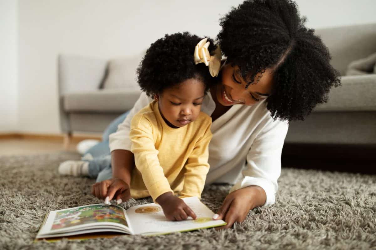 Parenthood concept. Adorable curly little african american girl playing with her mother, happy black toddler and mom reading book on floor, home interior, closeup, copy space