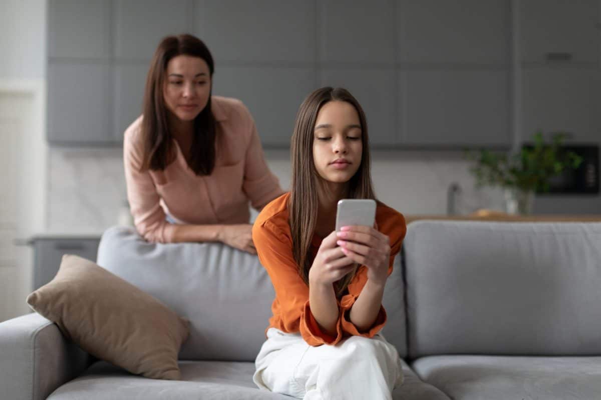 Parental monitoring of teens. Curious mother spying daughter, girl messaging on smartphone on sofa at home. Family and modern technologies