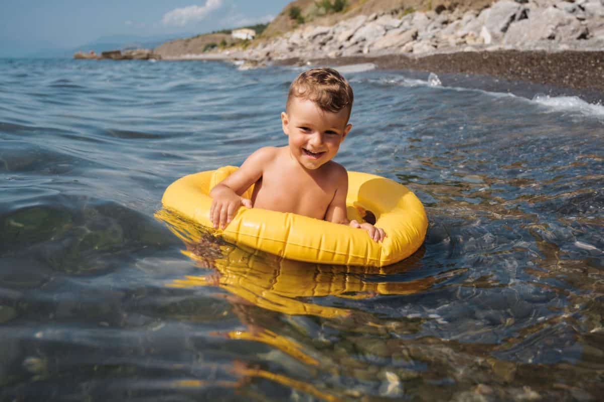 Baby boy swims with an inflatable yellow circle in the sea on a sunny day. The kid learns to swim and enjoys the game.