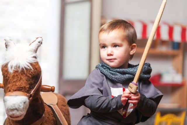 Toddler playing with sword next to rocking horse in playroom. Home play, symbolic play.