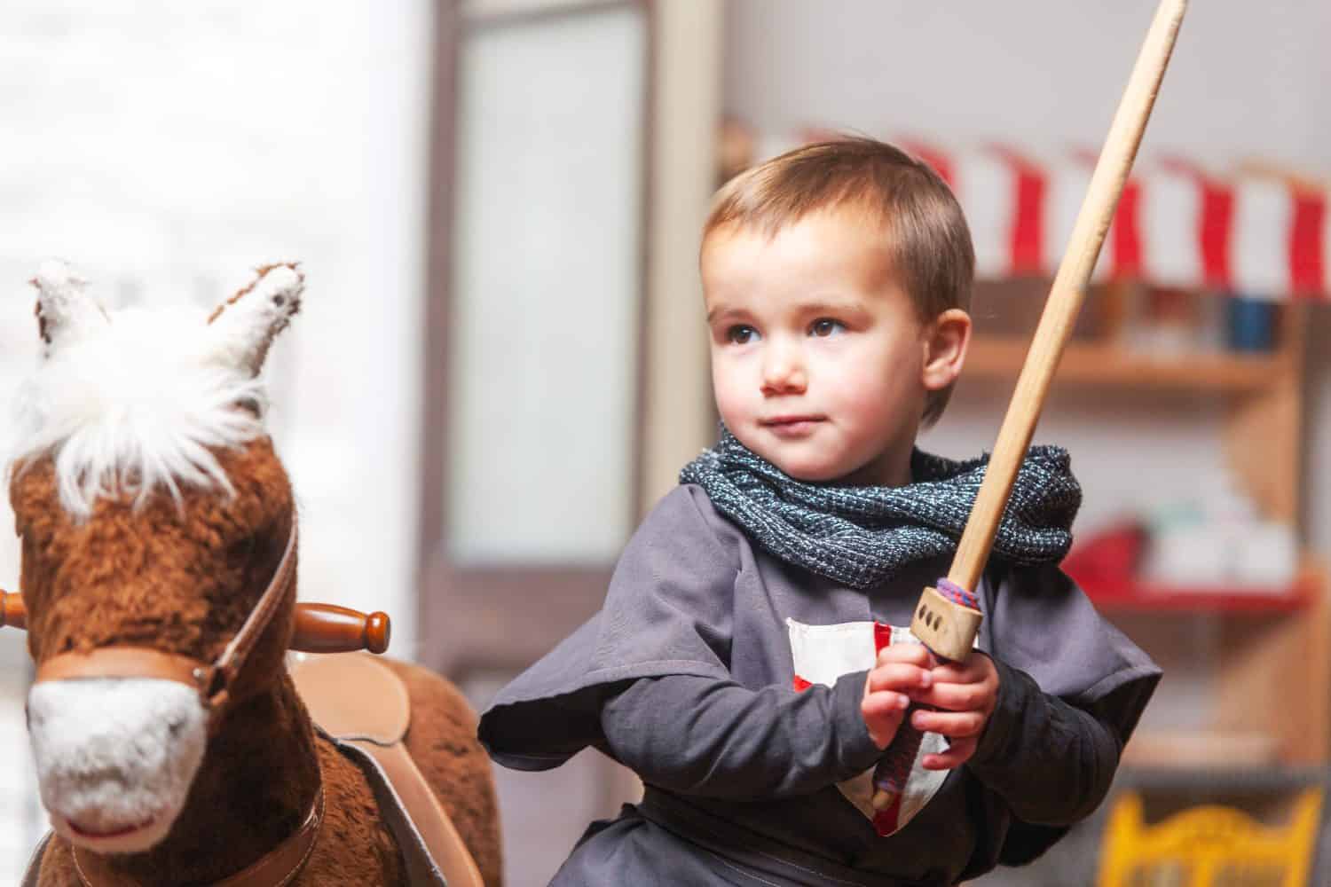 Toddler playing with sword next to rocking horse in playroom. Home play, symbolic play.