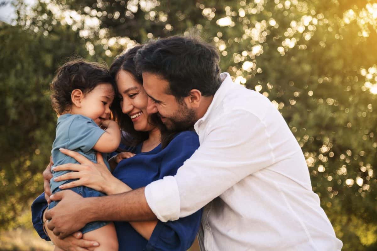 A joyful family moment with a father, mother in a blue dress, and their one-year-old son in blue clothes, hugging and smiling in a grassy natural setting with a tree in the background