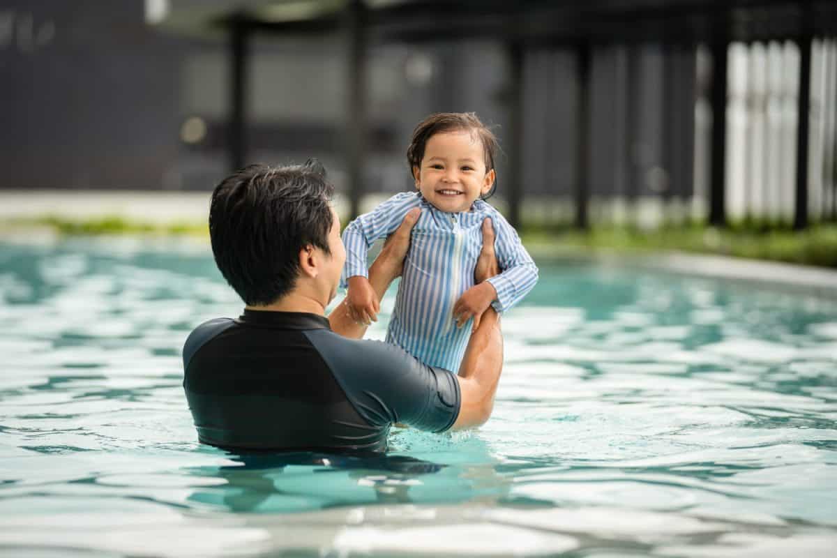 father holding and lifting todler baby in his arms while playing water in a swimming pool