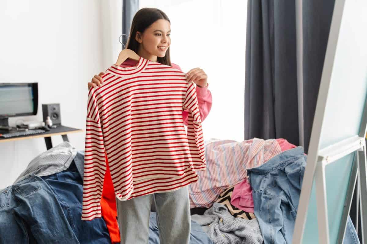 Young girl, teenager smiling while holding a red and white striped shirt on a hanger in her messy bedroom, deciding what to wear