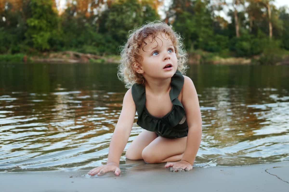 Fair-haired curly Caucasian little blonde girl dressed in polka dot swimming suit sitting by the water playing with sand while wading in warm summer river