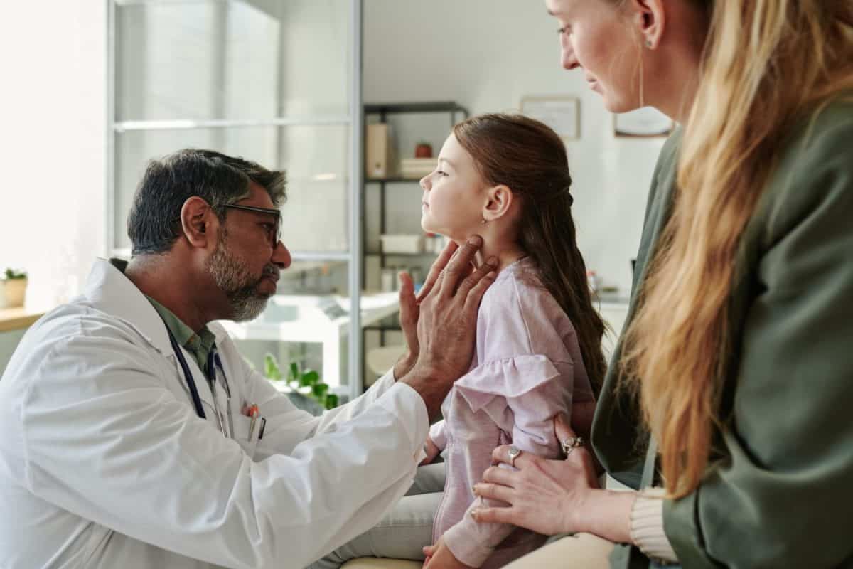 Side view of cute little patient of endocrinologist having her thyroid checked by experienced male clinician during consultation