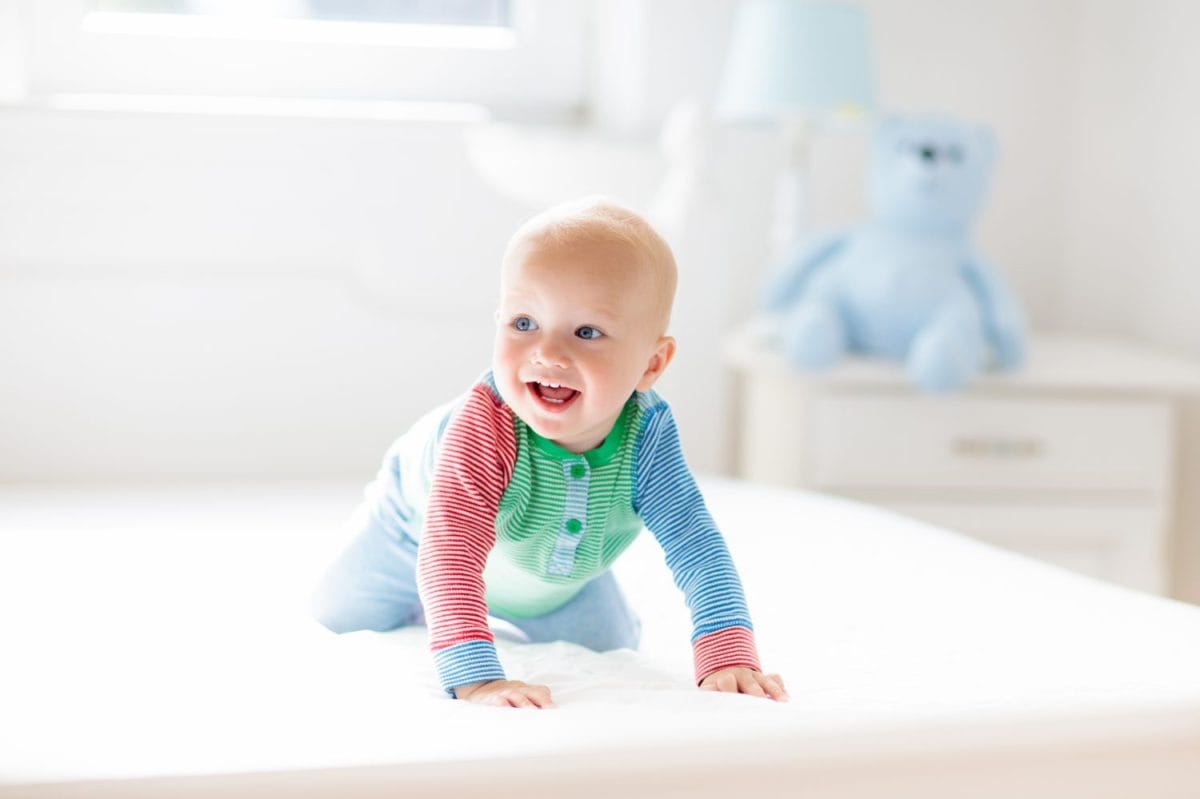 Baby boy crawling on bed. Little child playing in white sunny bedroom. Infant kid learning to crawl. Nursery for children. Textile, clothing and bedding for kids. Family morning at home.