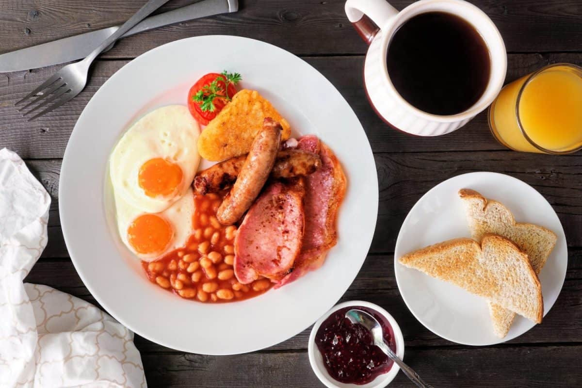 Full English breakfast with baked beans, bacon, eggs, sausage and hash browns. Overhead view table scene on a dark wood background. British Household Routines That Confuse Americans