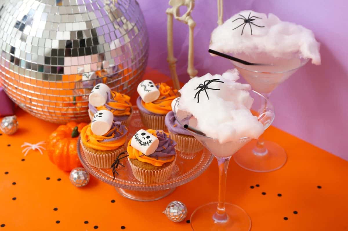 Halloween party composition with cupcakes, disco ball, drinks and decorations on orange table against purple background