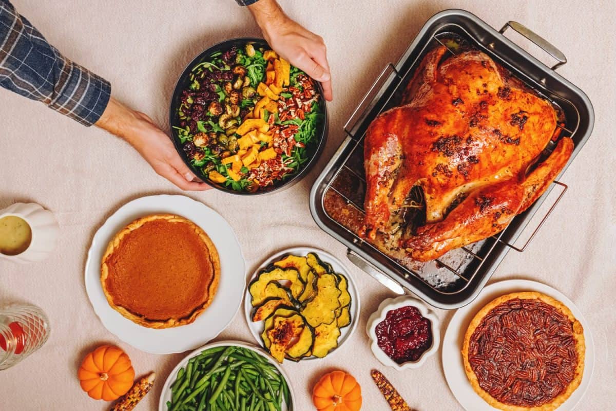 Man putting fresh salad on the table full of traditional Thanksgiving dishes with a golden-brown roast turkey, green beans, roasted squash and and pies