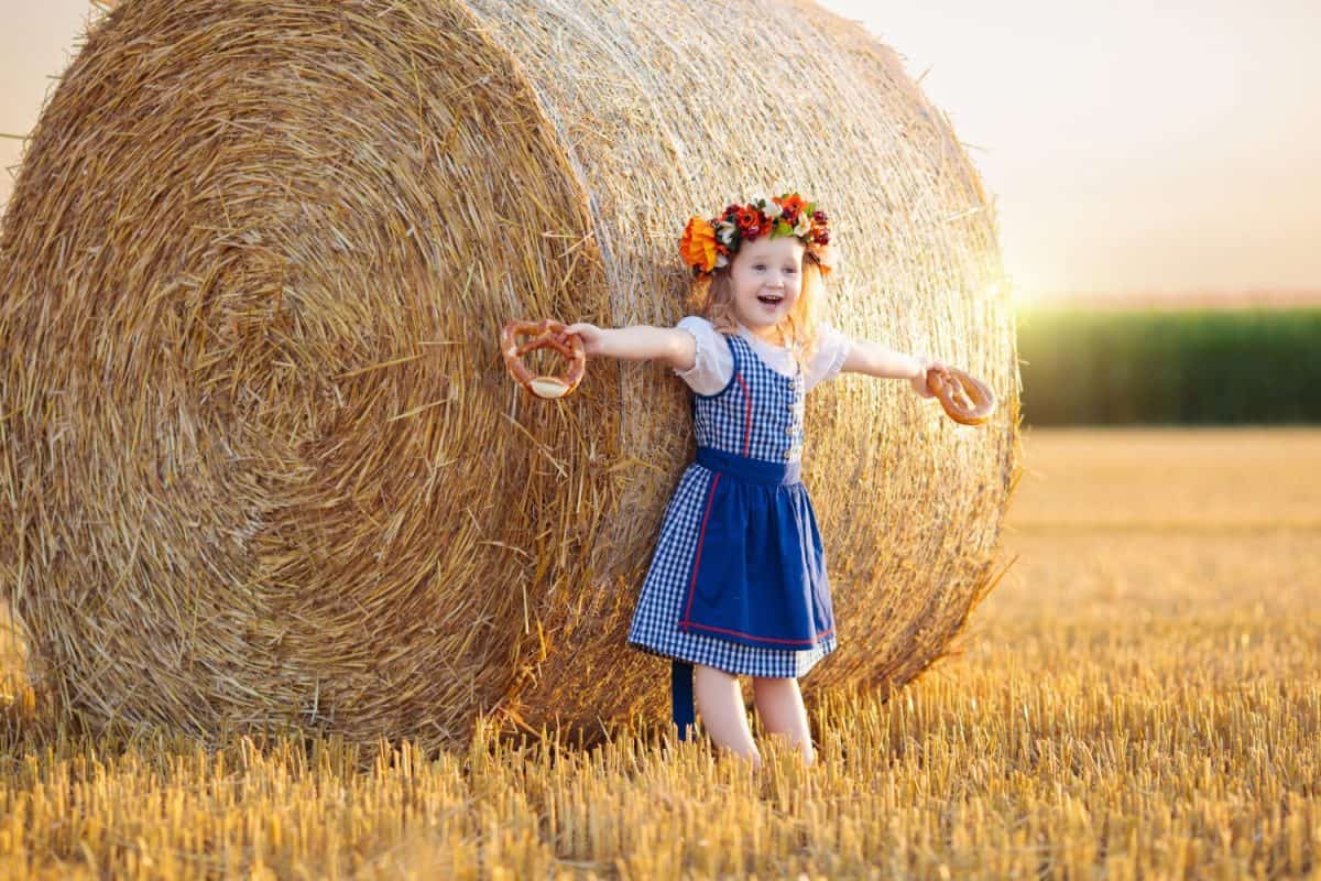 Child in wheat field with bread and pretzel. Oktoberfest time in Germany. Kids in traditional German dirndl dress and leather pants in autumn harvest crop field.