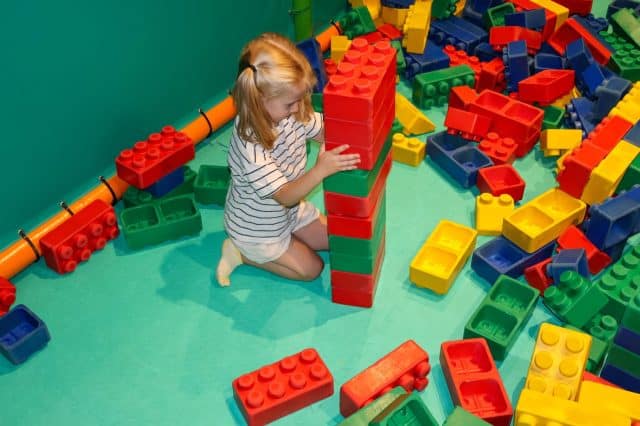 Little girl, toddler with builds a tower with a large multi-colored construction set. Play area for children. Large multi-colored construction set for children in the playroom. Montessori toys.