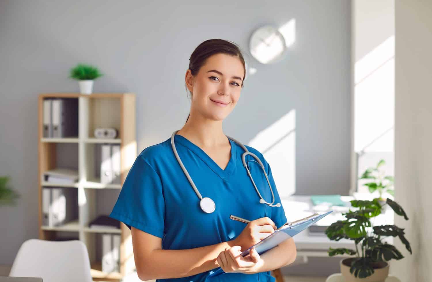 Portrait of happy female nurse or doctor at work. Beautiful young woman clinical nurse or physician in medical uniform holding pen and clipboard, standing in office, looking at camera and smiling