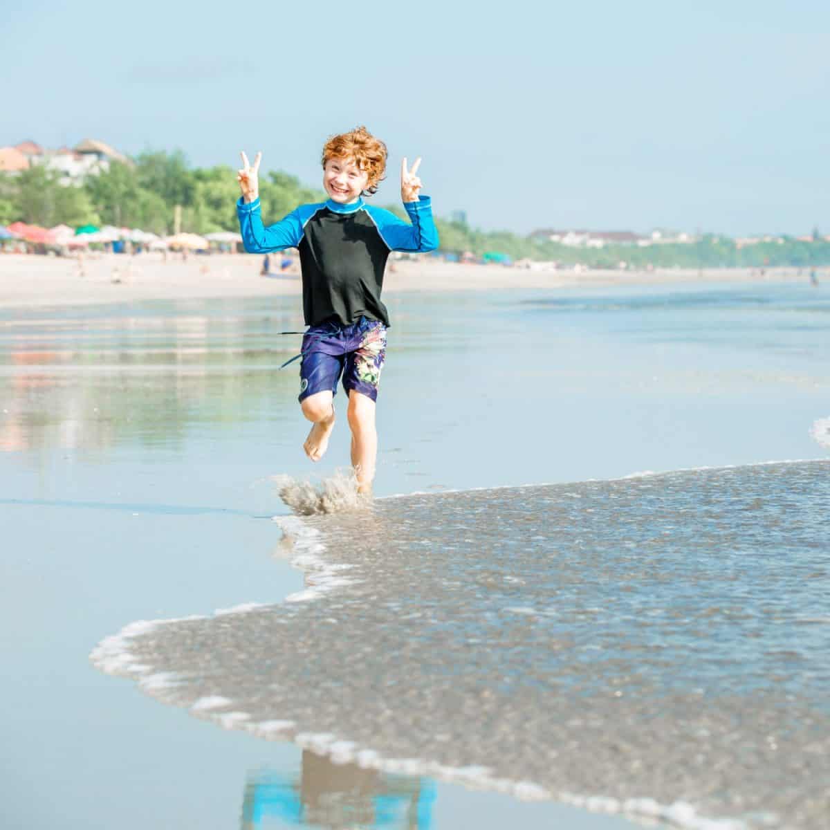 Young boy with red hair in swimming shorts and rushwest runs along Bali beach near sunset with reflection in the water