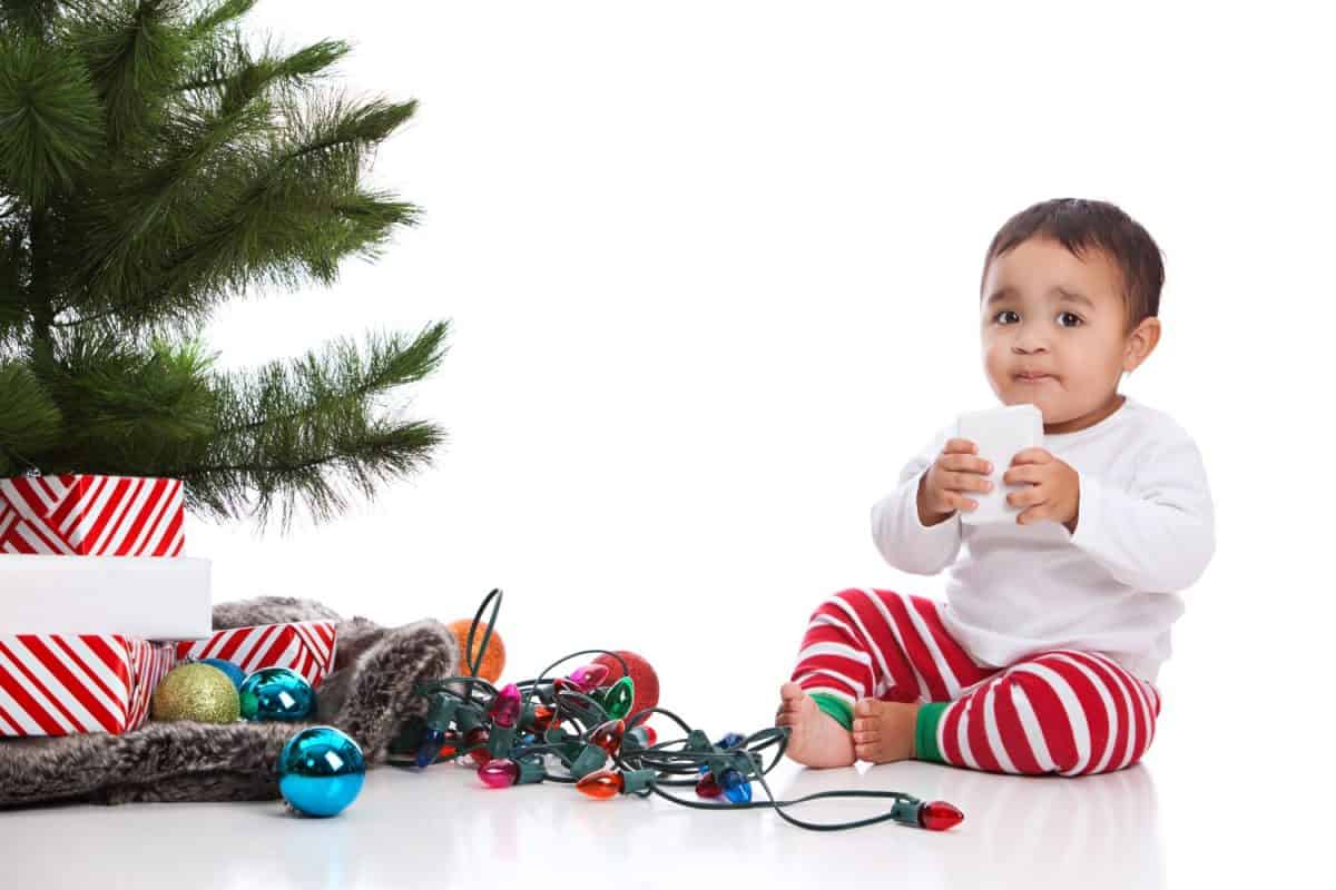 Christmas! Adorable mixed race baby boy wearing pajamas and sitting next to the Christmas tree with presents, lights and Christmas bulbs. Isolated on white.