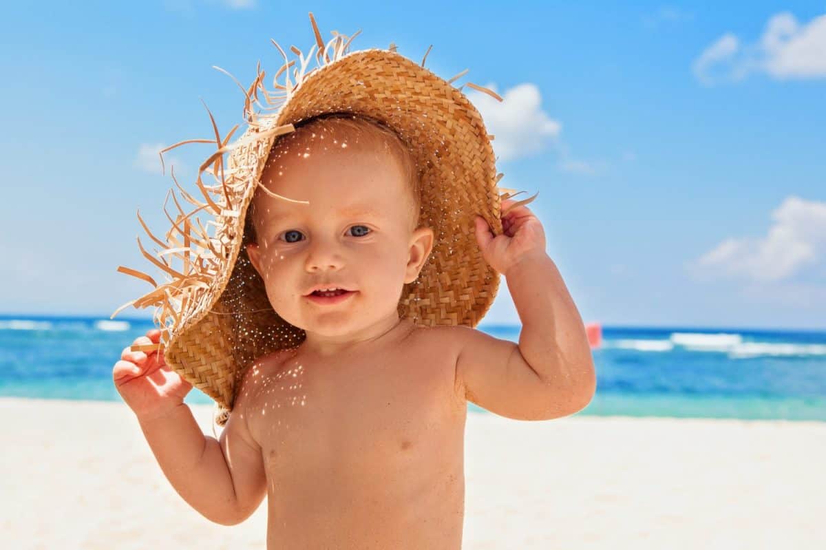 Funny photo of happy baby boy on beach with straw hat and dirty face covered with sand. Family travel, healthy lifestyle, recreation, water outdoor activity on summer beach vacation with children.