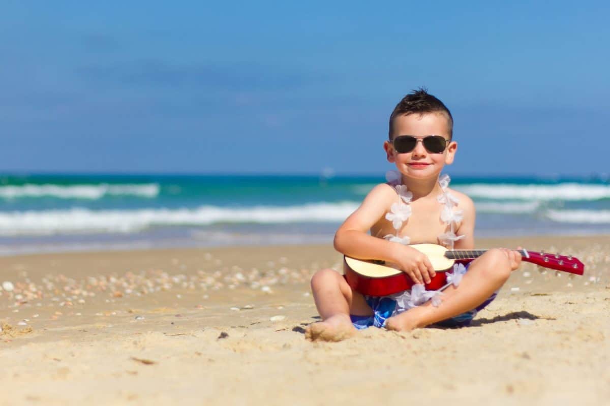 Little happy boy have fun, play music on Hawaiian guitar , enjoying sun ocean beach. Children healthy lifestyle. Travel, family activity on tropical island summer holiday