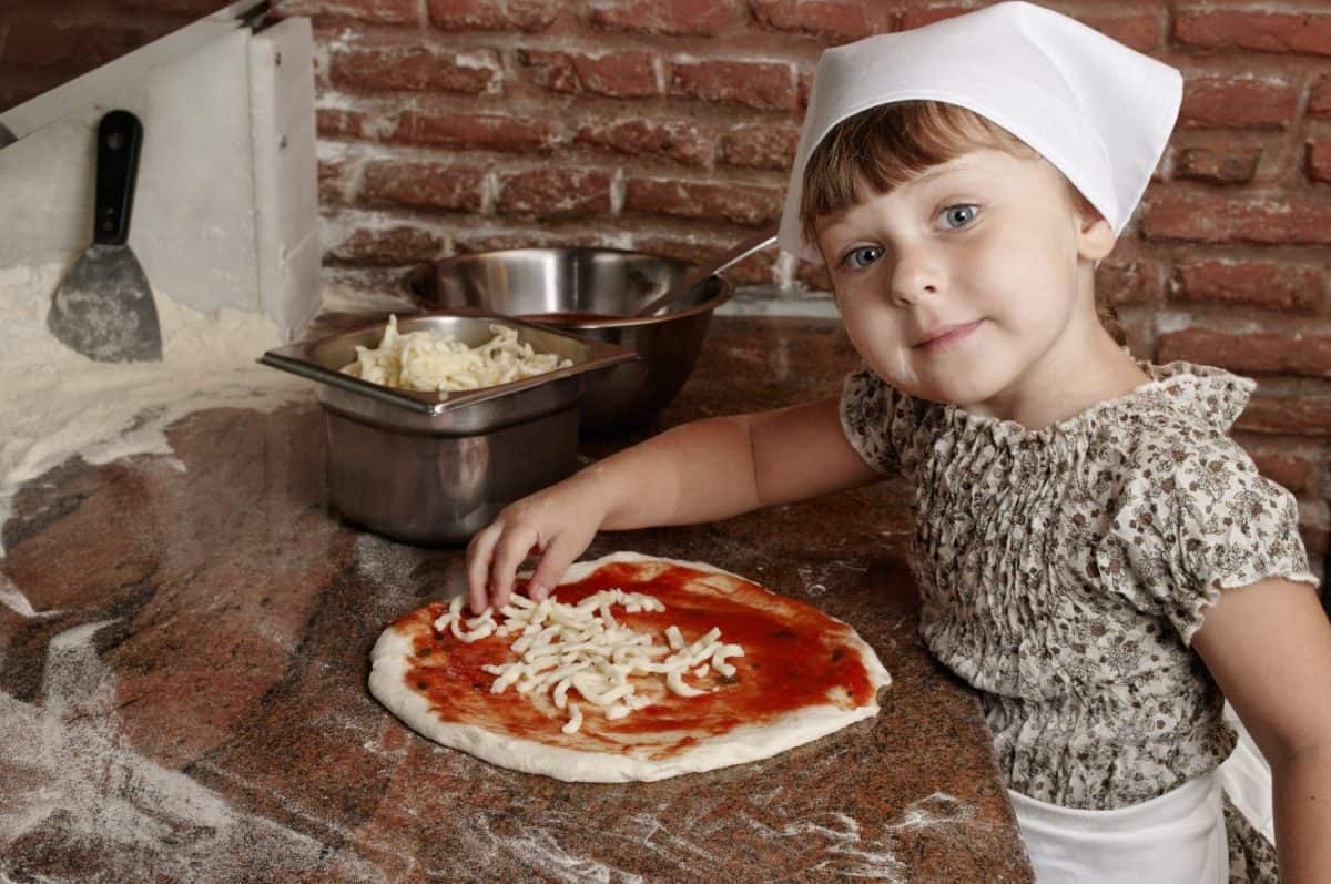 Little girl preparing pizza on the professional kitchen (old italian pizzeria). Adding cheese to pizza