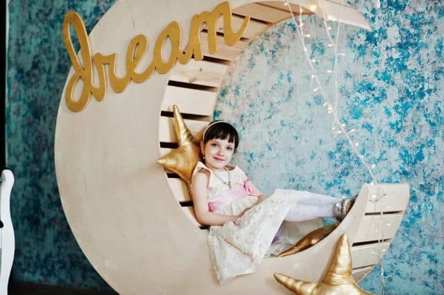 Beautiful little girl in a dress posing next to wooden moon decoration with golden star pillows.