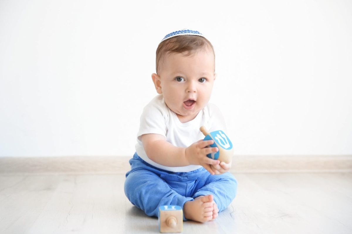 Cute baby in kippah playing with dreidels while sitting on floor at home