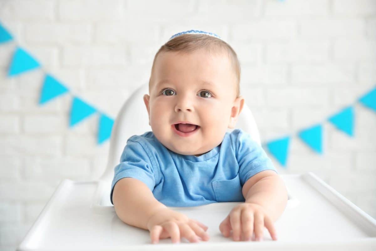 Cute baby in kippah sitting on high chair against brick wall