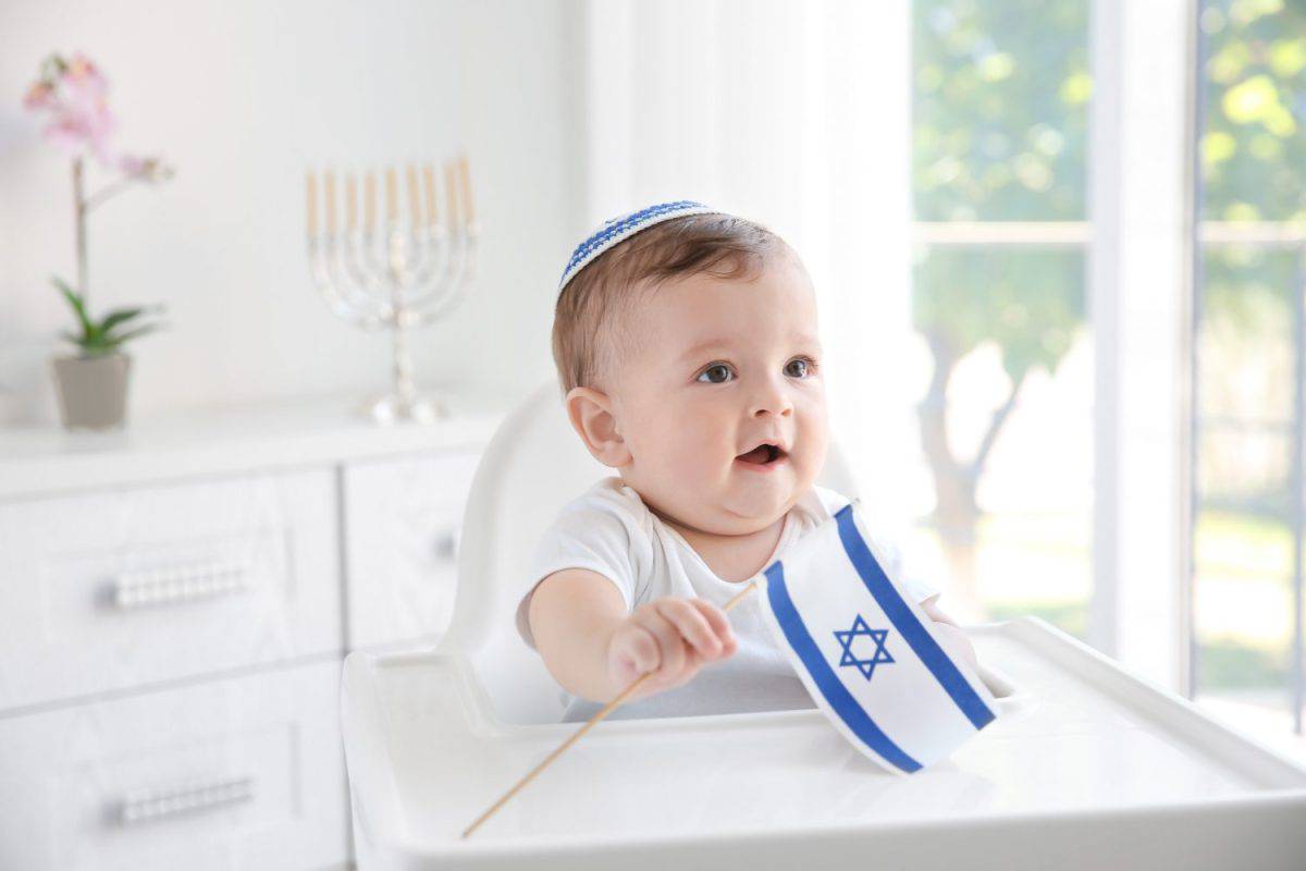 Cute baby in kippah with flag of Israel sitting on high chair at home