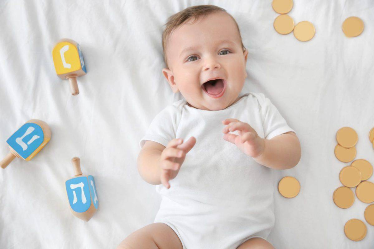 Cute funny baby lying near dreidels and chocolate coins on bed