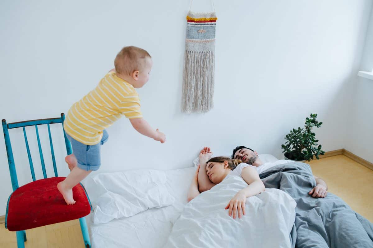 Cute little toddler son is jumping on bed from chair. He's determined to wake his parents, sleeping there in the morning.