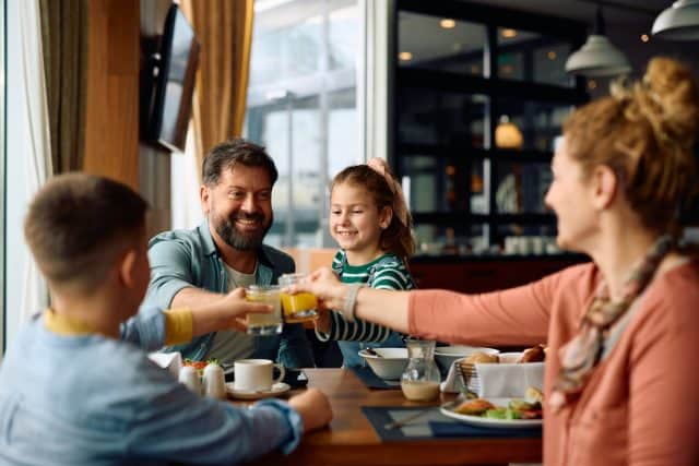 Happy family toasting while eating breakfast in a restaurant during their hotel stay.