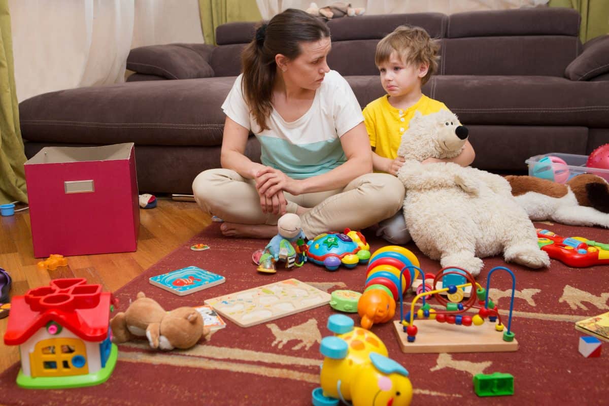 Young pregnant mother and her little son, at home. Mother and little son sit on a floor. Mom tired to tidy up the house. Child scattered toys. Mess in the house