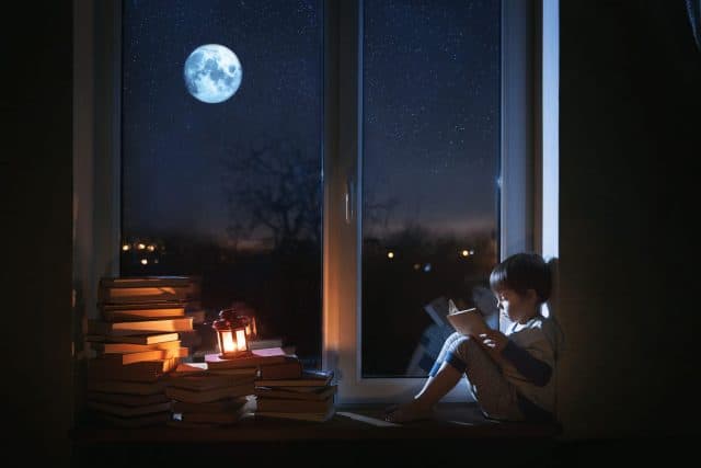 A cute boy sits on the windowsill at night. The child reads books under the moonlight. The window shows the moon and stars.