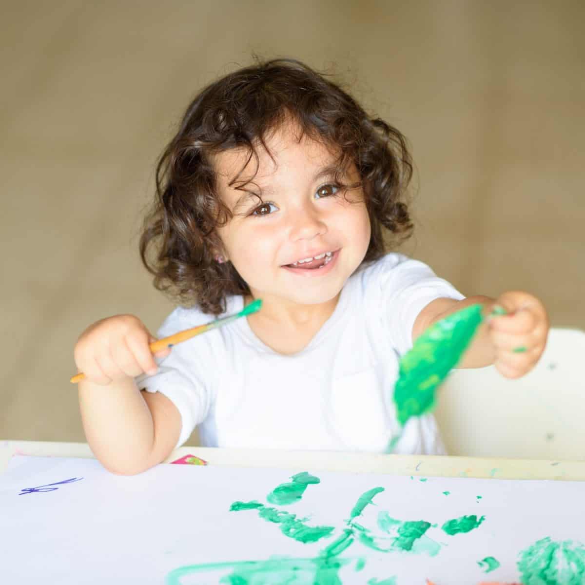 Cute little girl drawing with green paint on fall lives. Curly adorable child painting at table.