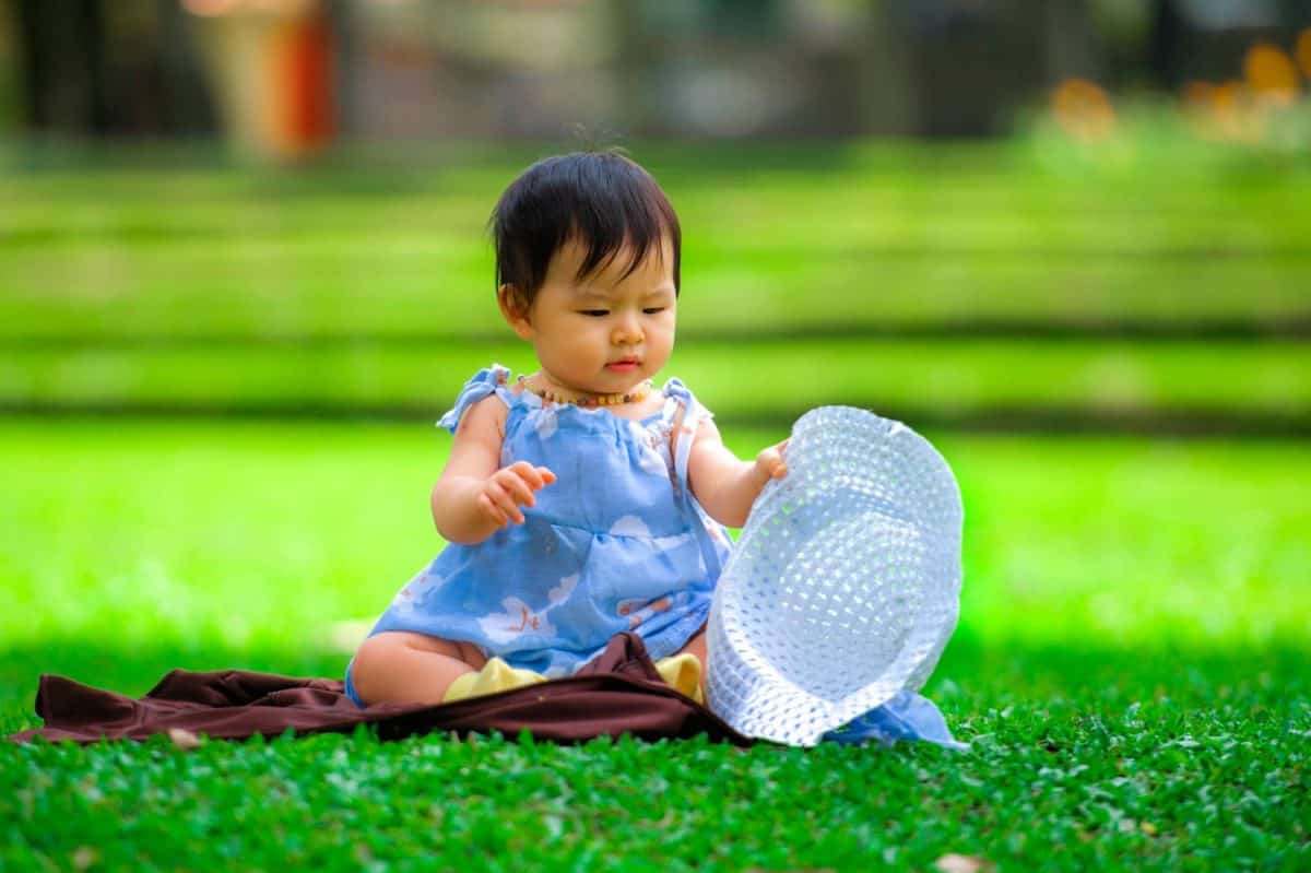 isolated candid portrait of sweet and adorable Asian Japanese baby girl 3 or 4 months old playing with hat alone at city park sitting on green grass in childhood innocence and purity concept