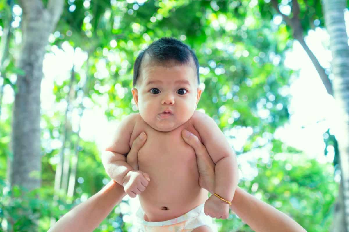 Southeast Asian new born isn't wearing shirt. South East Asian newborn is 3 months old. Mother's holding Infant with hands. Back side of young kid is trees. Background of child is green & freshness.