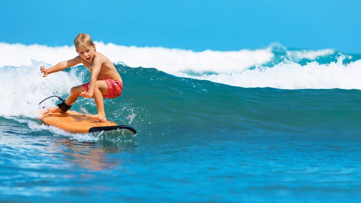 Happy baby boy - young surfer learn to ride on surfboard with fun on sea waves. Active family lifestyle, kids outdoor water sport lessons, swimming activity in surf camp. Summer vacation with child.