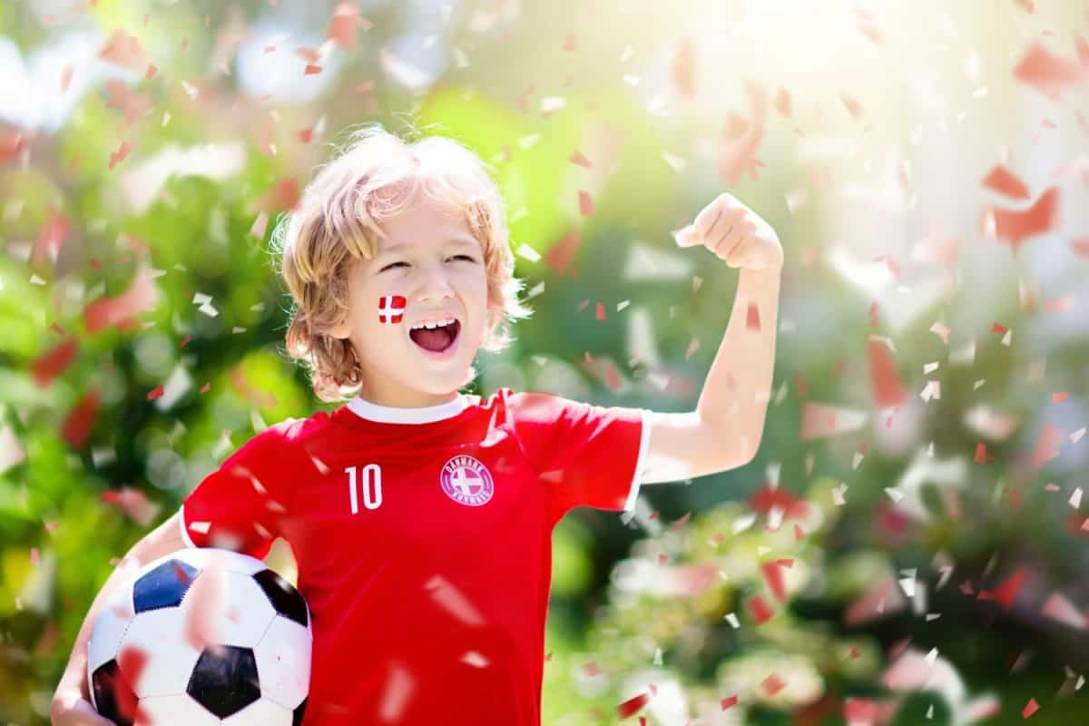 Denmark football fan cheering. Danish kids play soccer and celebrate victory on outdoor field. Danmark team supporter. Little boy in Dansk flag jersey kicking ball. Sports training for children.