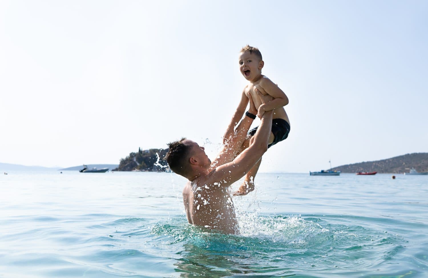 Dad and son are playing in the sea. The father tosses the son up. Greek islands, water splashes, vacations, blue sky and sunny day