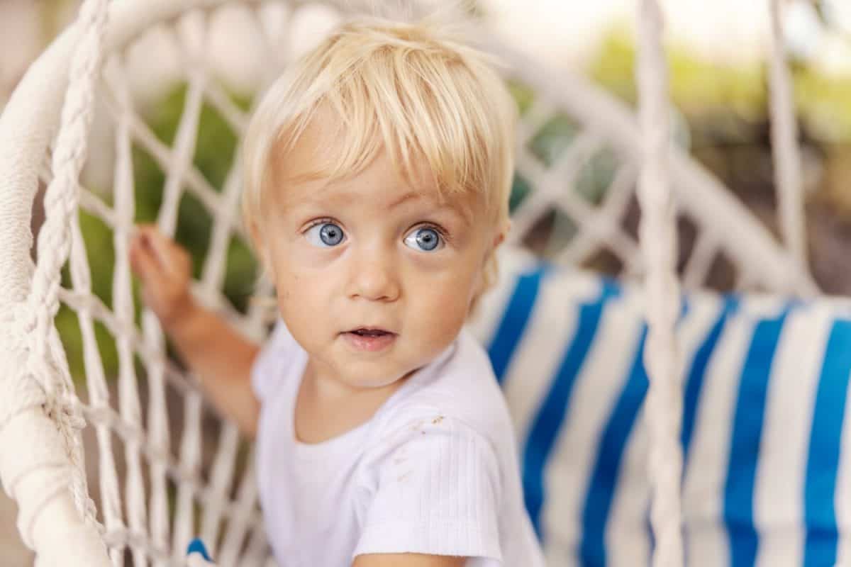 Too cute kid, baby's face is in focus. A toddler in a white bodysuit for children with beautiful blue eyes and blond hair looked sideways thoughtfully. Innocent children smile and pure happiness