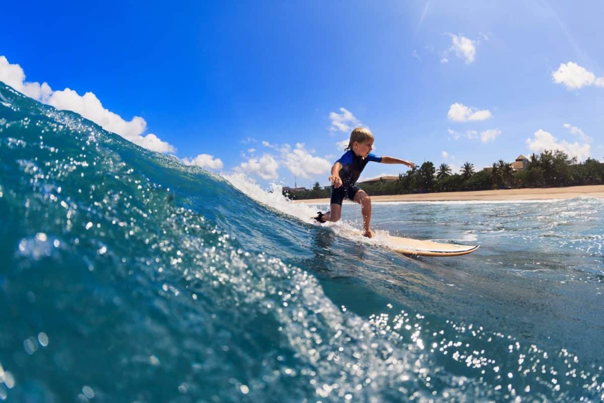 Happy surf boy - young surfer learn to ride on surfboard with fun on sea waves. Active family lifestyle, kids outdoor water sport lessons, swimming activity in surfing camp. Summer vacation with child