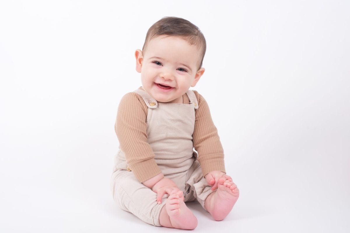 Adorable baby boy wearing beige overalls sitting on white background looking at camera and smiling.