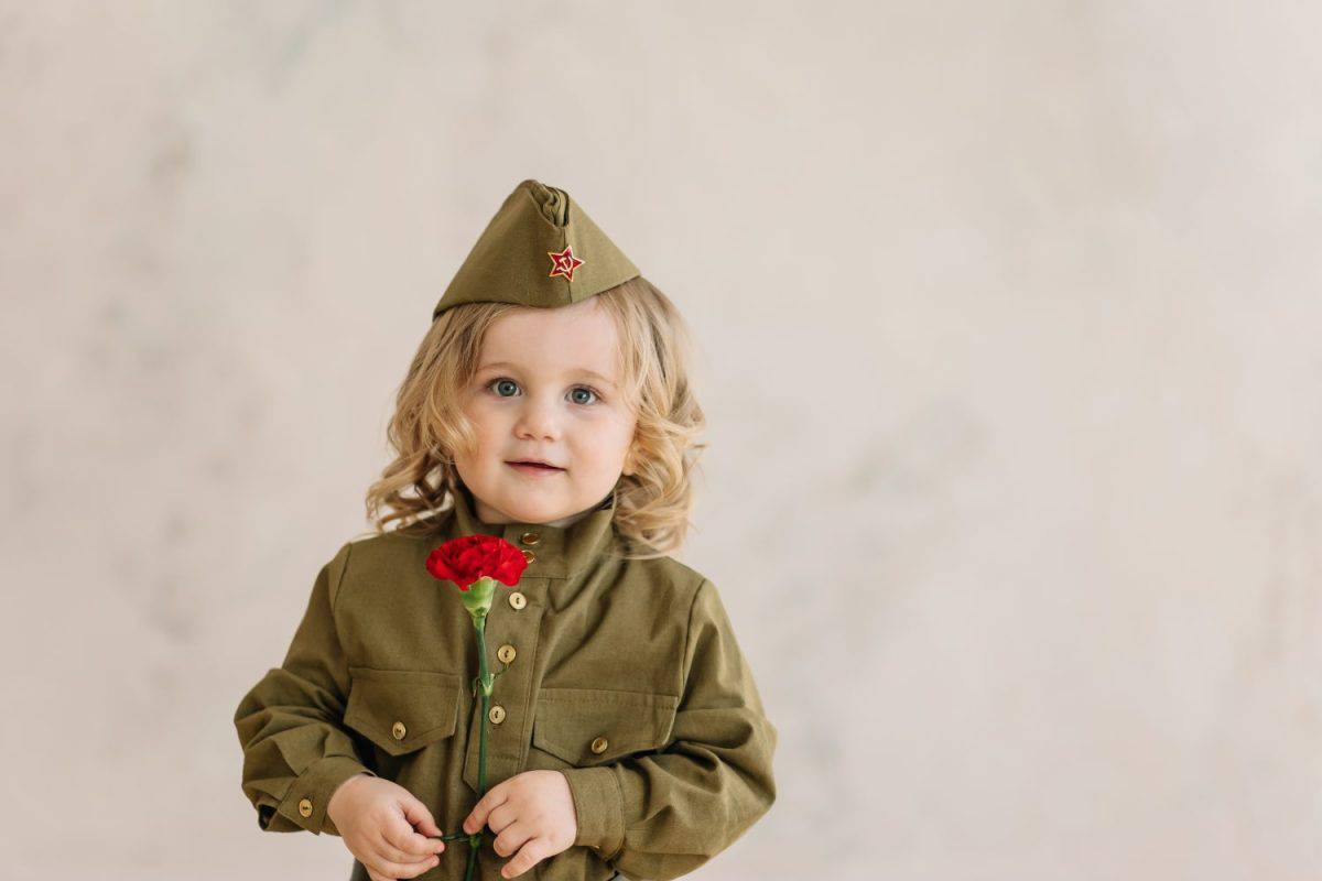 a nice curly baby girl in the old fashioned military uniform of the red army and forage-cap with red carnations on beige background. Victory day 9 May. postcard for a veteran