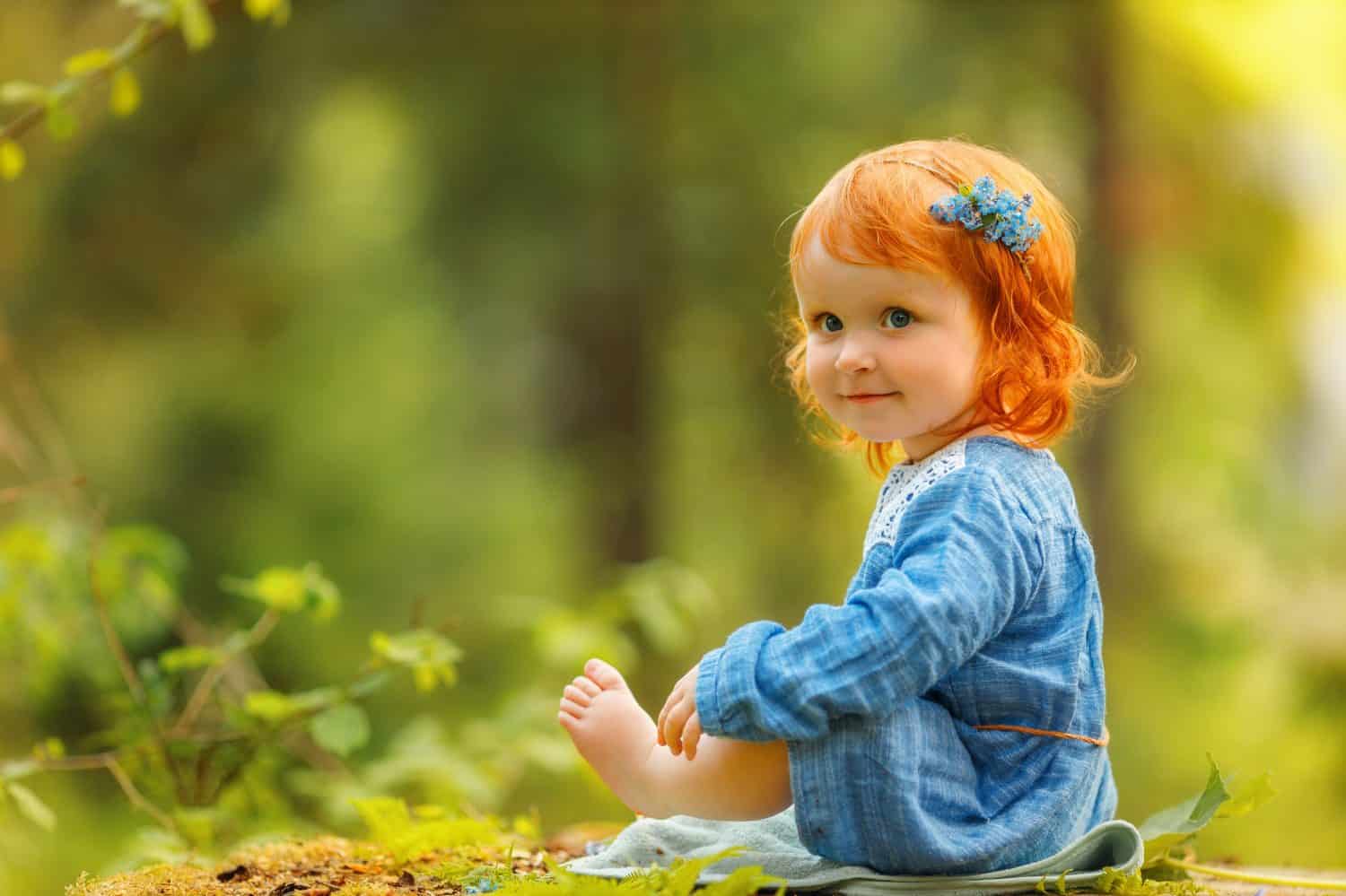 Baby red hair girl in blue dress sitting on a big tree stump in the park, looking at something invisible during sunset. Fantasy concept.