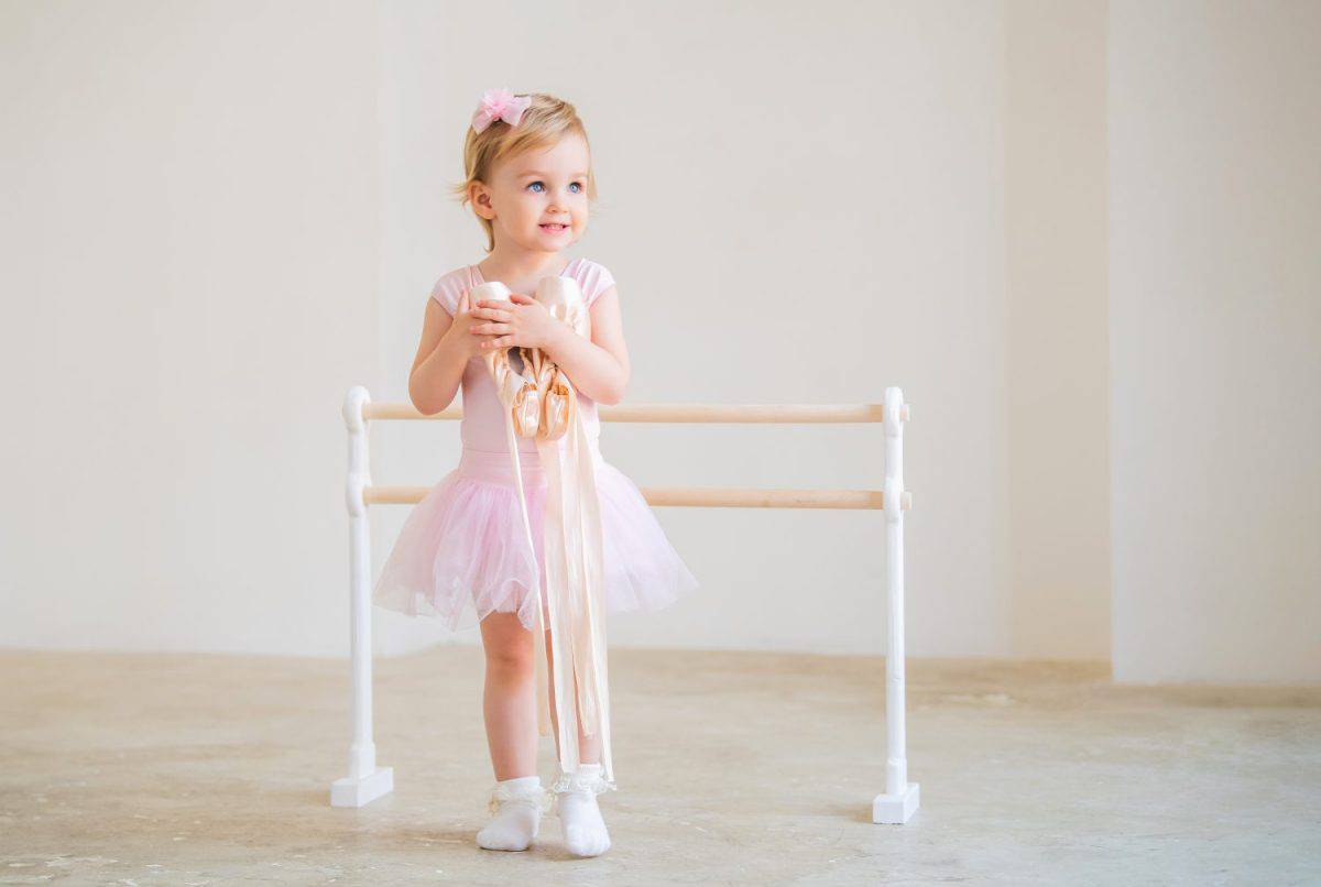 Portrait of cute blue-eyed baby ballerina in pink hugging pointe shoes.